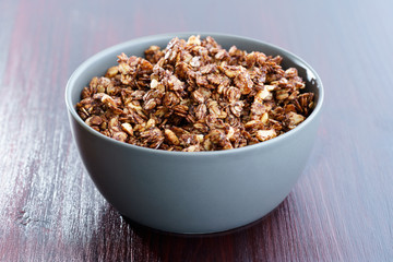 Homemade cocoa granola with crunchy peanuts and maple syrup in grey ceramic bowl. Morning light, red wooden table, high resolution