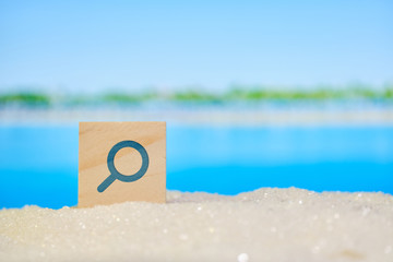 Magnifying glass on a wooden block in abstract sand with blurry photo of sea and beach. Travel and tour search concept.