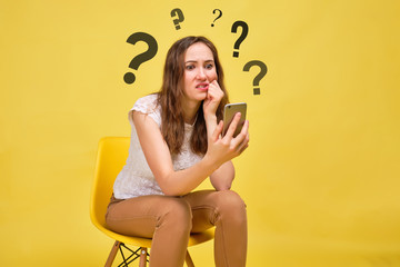 A girl in summer clothes on a yellow background sits and looks thoughtfully into a smartphone with questions above her head.