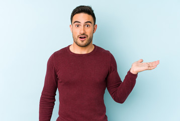 Young caucasian man isolated on blue background impressed holding copy space on palm.