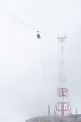 A cable car, mountain gondola suspended on a rope disappearing in the fog. The red-white pylon steel construction is barely visible.