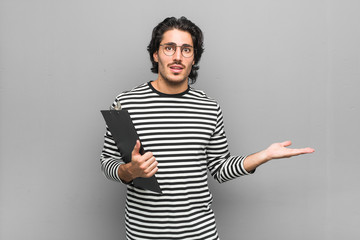 Young employee man holding an inventory shocked and amazed holding a copy space between hands.