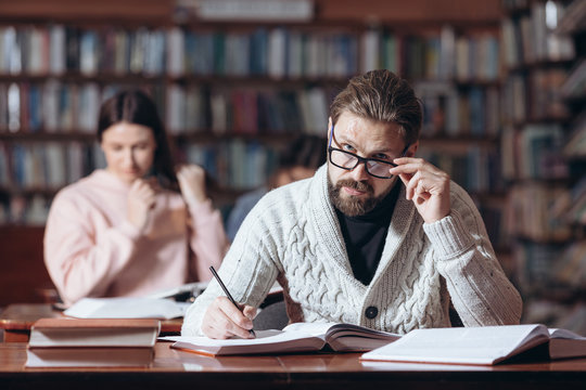 Confident middle-aged man in knitted sweater and eyeglasses taking notes while reading books at library. Bearded reader looking at camera while sitting at table with books.