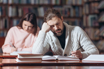 Tired bearded man in black polo neck and knitted sweater fall asleep while searching necessary information at public university. Mature reader sleeping while sitting at table with open books.