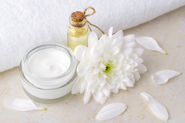 Face cream in a glass jar, bottle of chrysanthemum essential oil and white chrysanthemum near white terry towel on a table. Beauty, skincare and cosmetology.