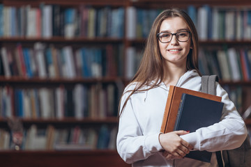 Portrait of happy young lady in white hoodie and eyeglasses holding books and looking at camera...
