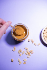 Woman's hand with a spoon taking peanut butter from a glass bottle on purple background. Vegan and bio snack. Vertical picture. Top view.