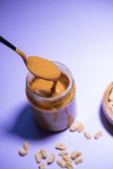Spoon with peanut butter falling to a glass bottle on purple background. Vegan and bio food. Vertical picture. Close up view.