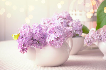 Room interior with purple lilacs flower blossom in kettle on table and yellow butterfly, tender romantic spring home decor in soft morning light, shiny glowing bokeh, decorating house with syringa