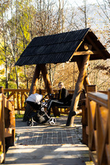 Happy mother at wooden Swing Seat In Garden with kid at carriage. Lovely garden swing in a beautiful landscape design. Swinging bench in the garden. Rest and maternity concept.