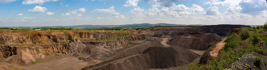 View of basalt (brown rock) quarry with rock exploitation during summer - blue sky with clouds, open-pit plant and village and background