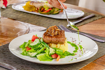 Grilled steak, caramelized onions, coffee sauce, mashed potatoes and vegetable salad on a white plate, hand holding a spoon and serving green mint sauce, blurred restaurant background