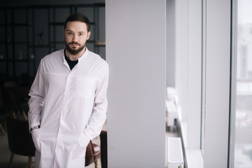 Naklejka na ściany i meble Confident young male doctor. Caucasian doctor with beard stands by the window and looking at camera. Male doctor posing in modern meeting office room of the hospital. Concept of medical work.