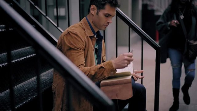 Latin male in stylish outfit writing information in personal planner while sitting at urban setting in city environment, trendy dressed student wearing backpack and walking at street