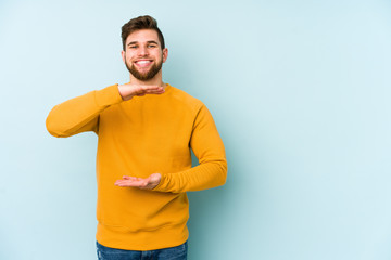 Naklejka premium Young caucasian man isolated on blue background holding something with both hands, product presentation.