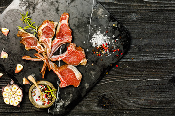Raw lamb chops with salt, pepper and dry herbs on dark stone plate on black old rustic wooden table, flat lay 