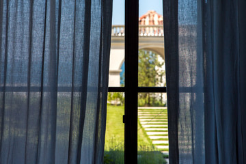 Window frame with blue curtains inside view of the backyard with green grass lit by sunlight on summer day.