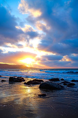 Obraz premium Dark wet big stones on the beach at the water edge at sunset. Marine landcape with heavy dark clouds and setting sun.