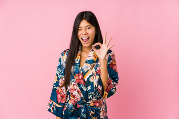 Young asian woman wearing a kimono pajamas winks an eye and holds an okay gesture with hand.