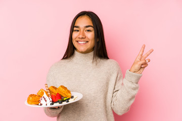 Young asian woman holding a waffle isolated joyful and carefree showing a peace symbol with fingers.