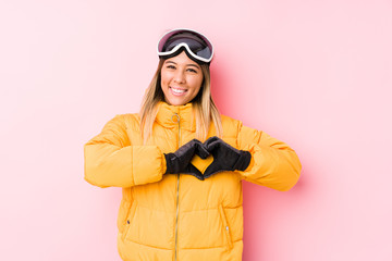 Young caucasian woman wearing a ski clothes in a pink background smiling and showing a heart shape with hands.