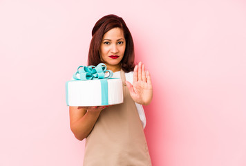 Middle age latin woman holding a cake isolated on a pink background standing with outstretched hand showing stop sign, preventing you.