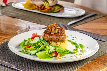 Grilled steak, caramelized onions, coffee sauce, and green mint sauce, mashed potatoes and vegetable salad on a white plate, , blurred restaurant background