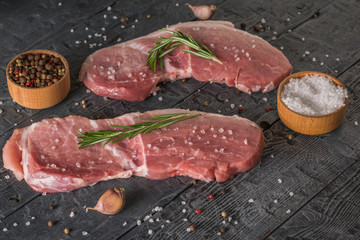 Two pork steaks with bowls of pepper and salt on a wooden table.
