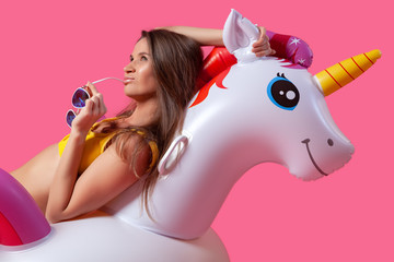 Summer Fashion  beautiful woman in summer clothes with sunglasses, smilling and posing  with balloons On Unicorn Float on isolated pink background