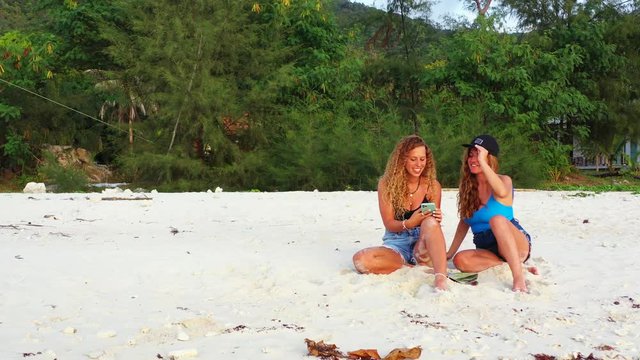 Two attractive young woman friends posing on camera taking selfies with smart phone with funny face expressions on white sand beach.