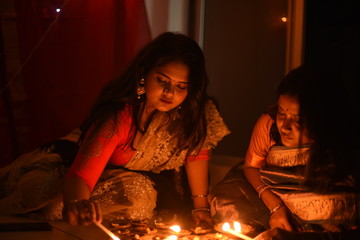 Two beautiful Indian Bengali women in Indian traditional dress are lighting Diwali diya/lamps sitting on the floor indoor in darkness on Diwali evening. Indian lifestyle and Diwali celebration