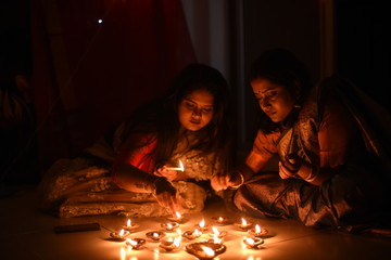 Two beautiful Indian Bengali women in Indian traditional dress are lighting Diwali diya/lamps sitting on the floor indoor in darkness on Diwali evening. Indian lifestyle and Diwali celebration