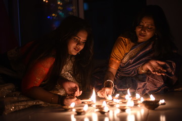 Obraz na płótnie Canvas Two beautiful Indian Bengali women in Indian traditional dress are lighting Diwali diya/lamps sitting on the floor indoor in darkness on Diwali evening. Indian lifestyle and Diwali celebration