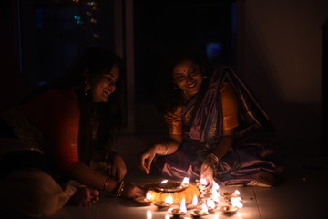 Two beautiful Indian Bengali women in Indian traditional dress are lighting Diwali diya/lamps sitting on the floor indoor in darkness on Diwali evening. Indian lifestyle and Diwali celebration
