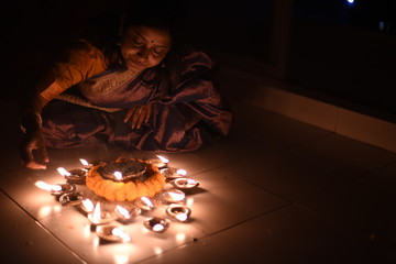 An young and beautiful Indian Bengali woman in Indian traditional dress is lighting Diwali diya/lamps sitting on the dark floor indoor to celebrate diwali. Indian lifestyle and Diwali celebration