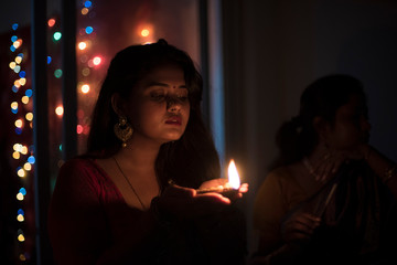 Obraz na płótnie Canvas Two young and beautiful Indian Bengali women in Indian traditional dress are celebrating Diwali with diya/lamp and fire crackers on a balcony in darkness. Indian lifestyle and Diwali celebration