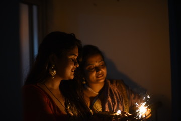 Two young and beautiful Indian Bengali women in Indian traditional dress are celebrating Diwali with diya/lamp and fire crackers on a balcony in darkness. Indian lifestyle and Diwali celebration