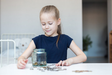 Cute little child girl counts and puts coins in a glass bottle in her room. The child saves money on the concept of the future