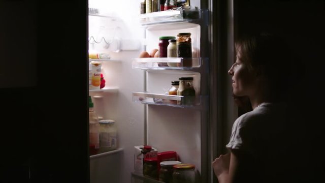 Young blonde woman opening the fridge in the dark. Taking pear. Sneaking for a snack during the night.