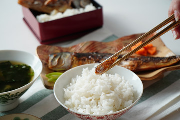 someone using chopsticks trying to pick a piece of grilled saba or mackerel fish served with cooked rice and miso soup on white and green striped placemat on white table