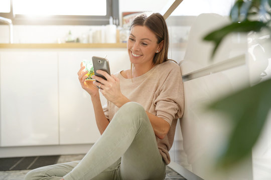 Woman sitting on kitchen floor, relaxing and using smartphone