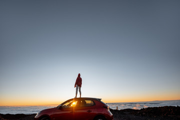Woman enjoying rocky landscapes above the clouds on a sunset, standing on the car highly in the mountains. Carefree lifestyle and travel concept