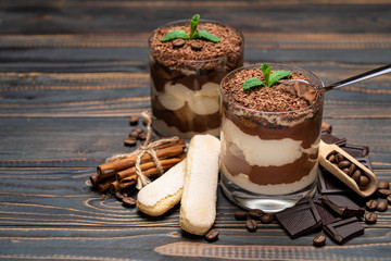 Classic tiramisu dessert in a glass cup, pieces of chocolate and savoiardi cookies on wooden background