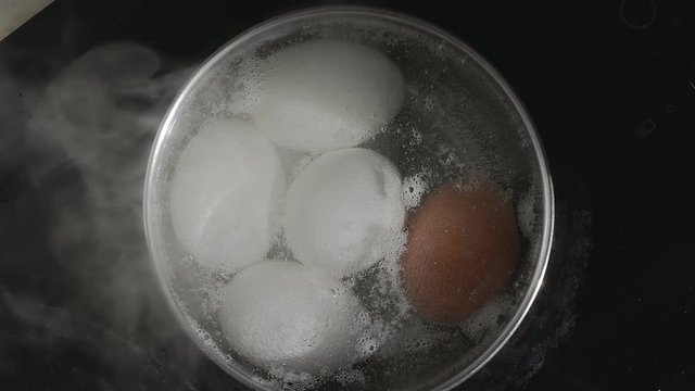 chicken eggs are cooked in a stainless steel pan. The water begins to boil and the eggs are covered with many small air bubbles. Sunlight from the window.