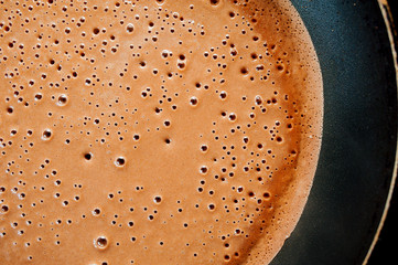 A pancake made of liquid chocolate dough with bubbles is fried in a black pan with drops of fat and...