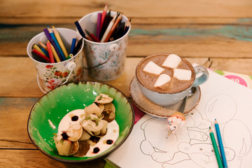 Small pancakes in a plate with berries and a Cup of cocoa with marshmallows, next to coloring with pencils, children's menu, selective focus