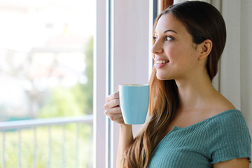 Close up of beautiful woman looking through the window drinking cup of tea at home, focus on model eyes, indoor photo