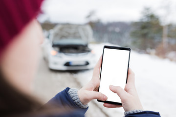 Close up of female holding blank screen smart phone against broken car. Winter scene.