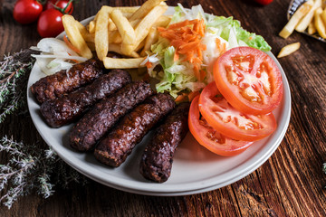 Spicy kofte (cevapcici, grilled minced meat) served with fresh tomatoes, lettuce, salad, vegetables and French fries on a rustic wooden board