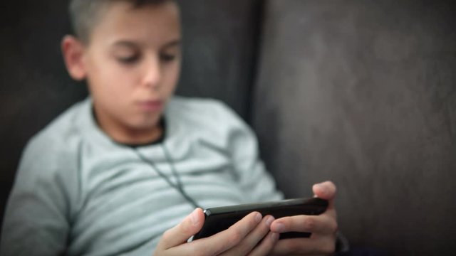 Little boy sitting on sofa during quarantine isolation with corona virus, playing with smartphone